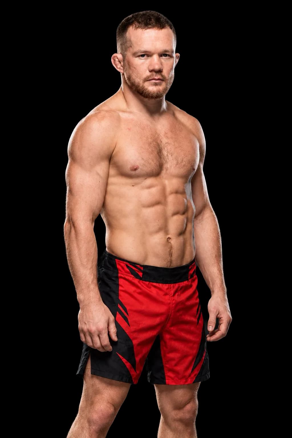 Petr Yan con torso descubierto y pantalón rojo y negro sobre un fondo gris oscuro.