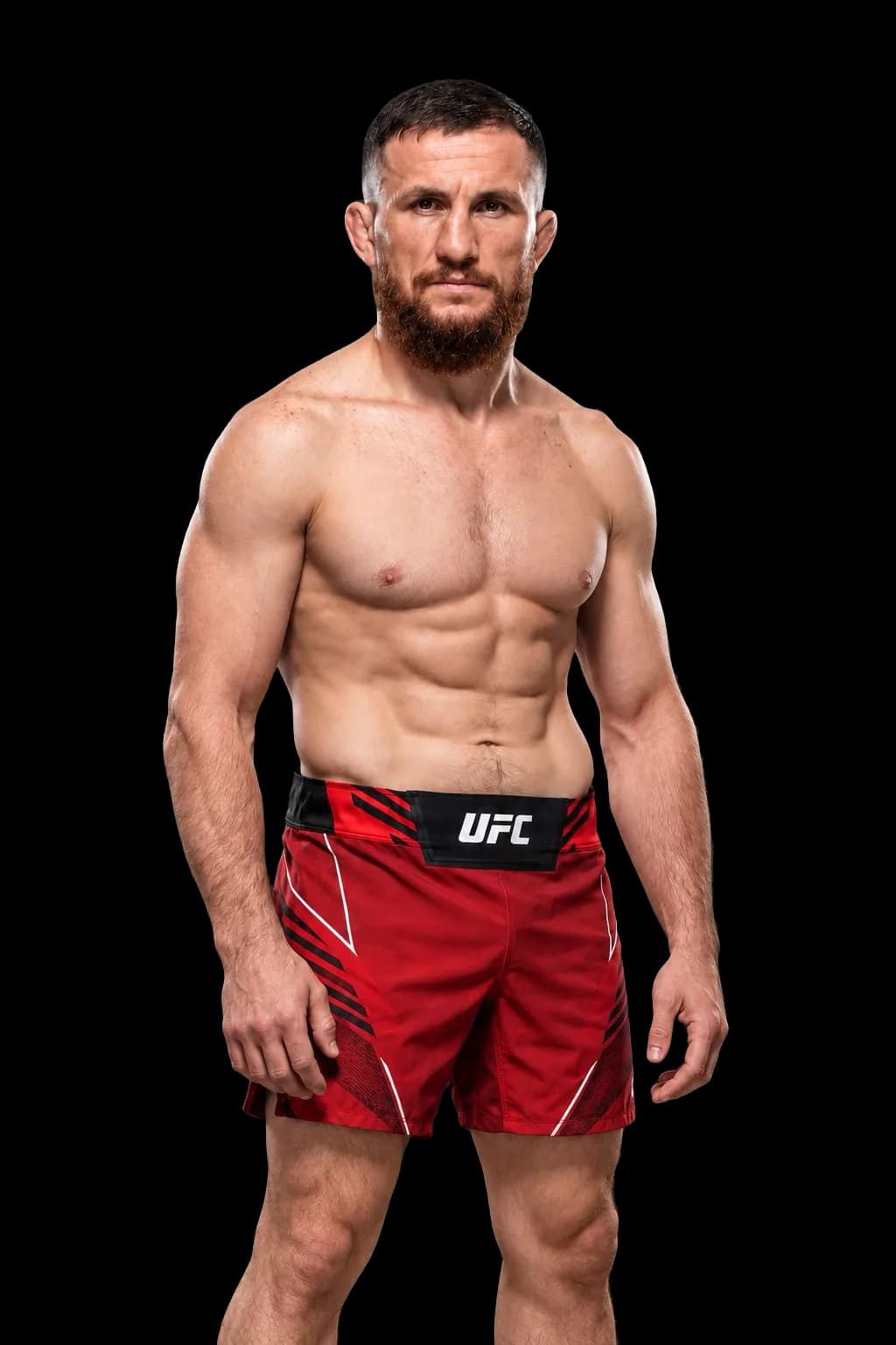 Merab Dvalishvili con torso descubierto y pantalón rojo sobre un fondo gris oscuro.